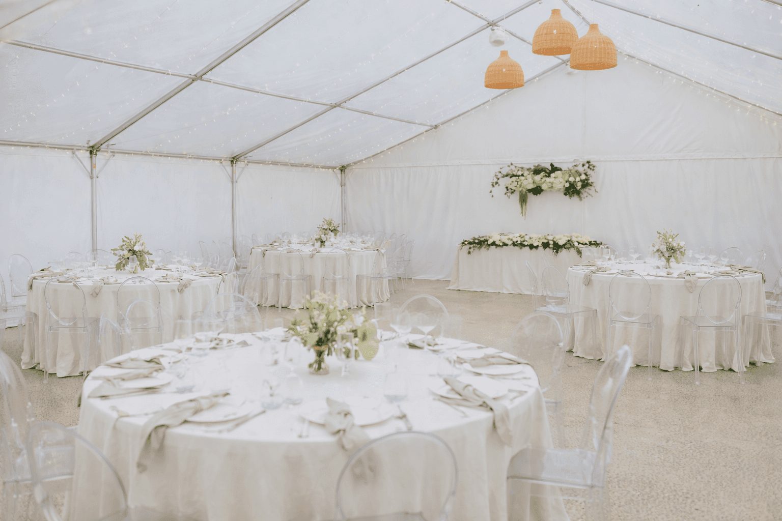 A white marquee set with round tables, white flowers and woven pendant lights